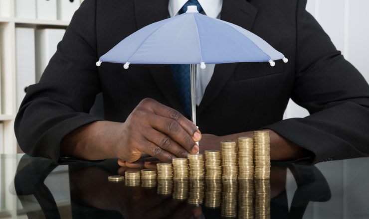 Close-up Of Businessman Protecting Stack Of Coins With Umbrella At Desk